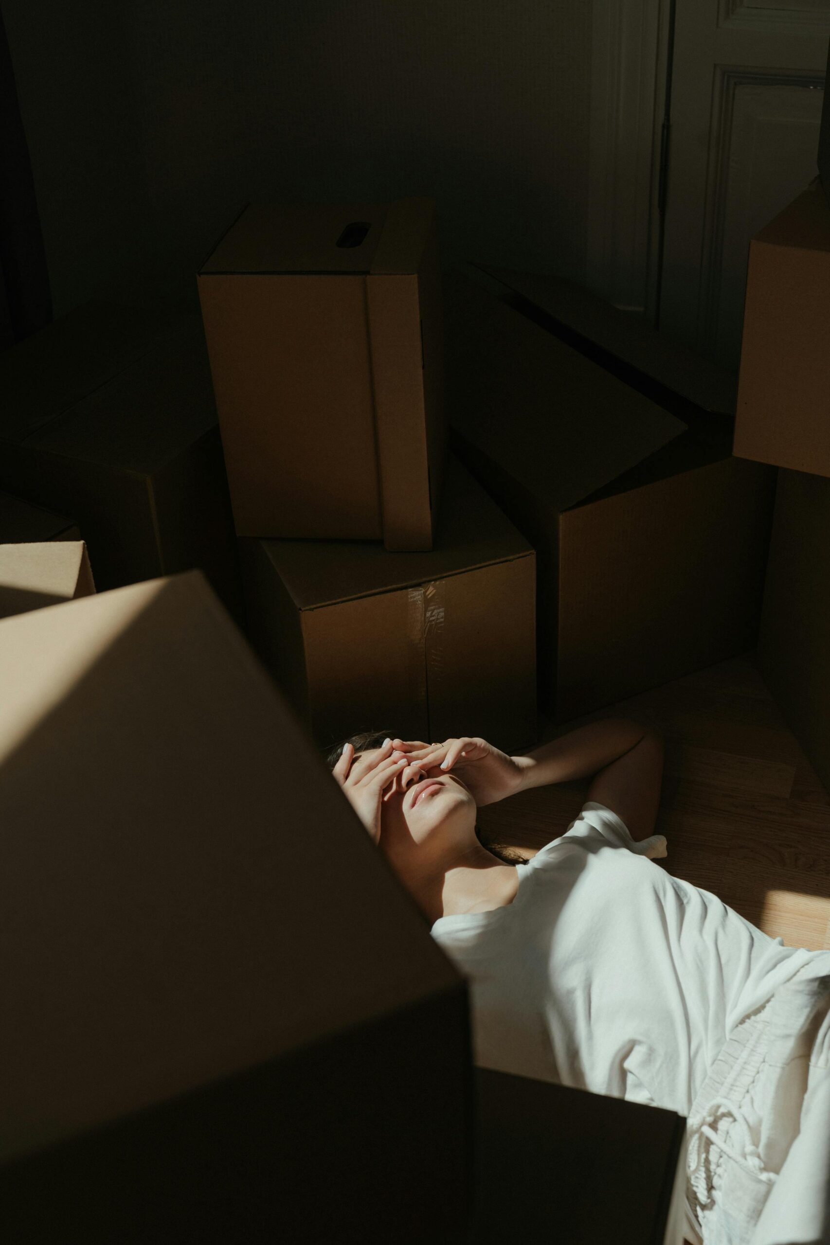 A woman lies on the floor surrounded by cardboard boxes in a dim sunlit room, expressing fatigue.